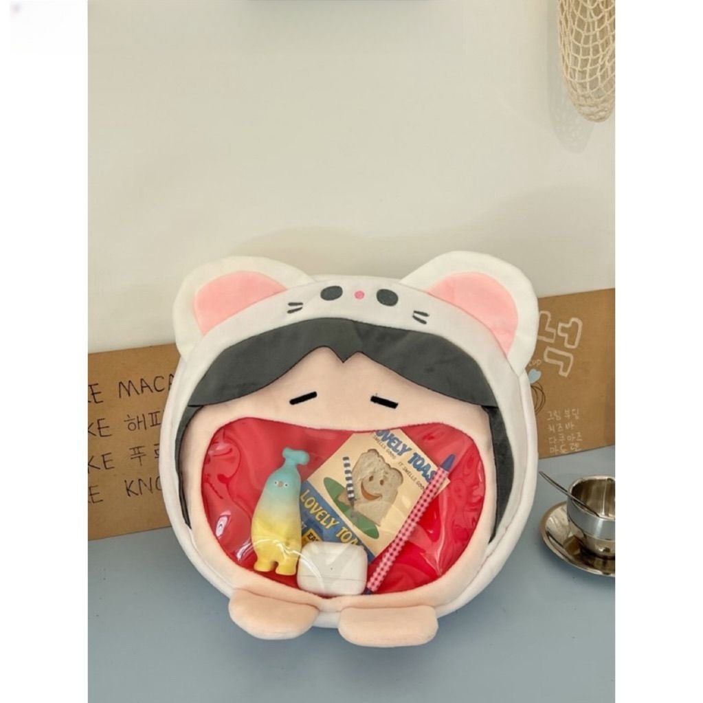 The Seakoff Adorable Cat Hoodie Plush Bag boasts a fun design with a transparent mouth pocket to hold items, including a packaged toy. Positioned on a table, it charms any room's décor with its cute accessories and whimsical appeal.