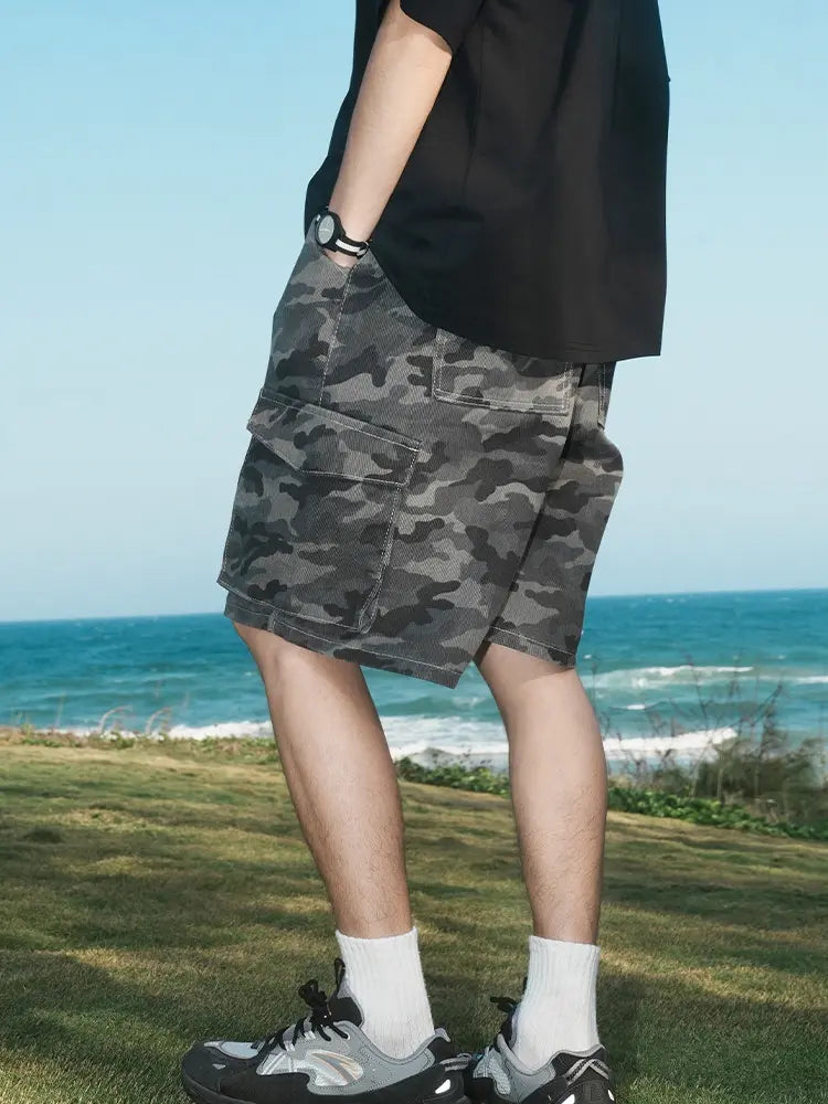 A person stands on grass near the ocean, wearing a black shirt, gray sneakers, white socks, a watch, and Men's Dark Grey Camo Cargo Shorts with Elastic Waist and Flap Pockets. Blue sky and ocean waves form the scenic backdrop.