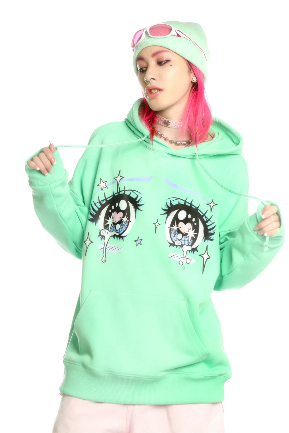 With pink hair, a person rocks Seakoff's mint green Kawaii Aesthetic Crying Eyes Hoodie, featuring large eye illustrations. They complete their anime streetwear look with a matching beanie, pink goggles, light-colored pants, and a choker while holding the hood's drawstrings.