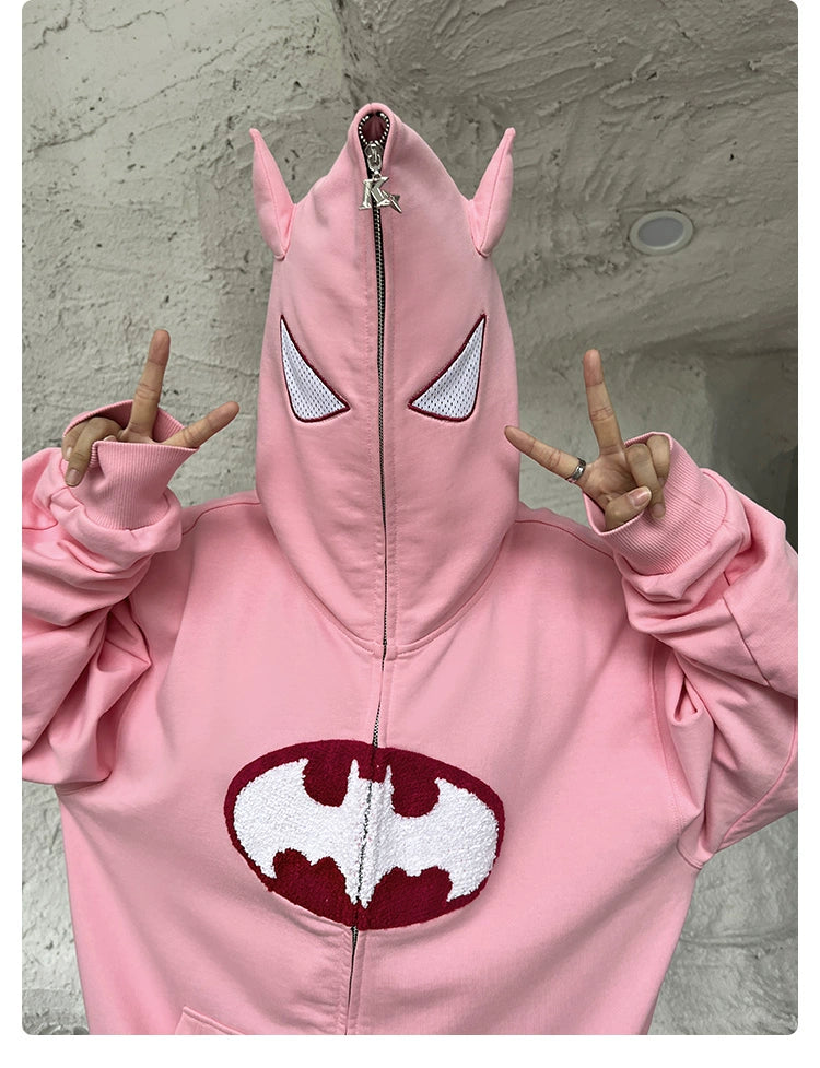 A person in a Seakoff Batman-Inspired Hoodie, a pink 400GSM oversized zip-up sweatshirt with white mesh eyes and small ears, making peace signs with both hands. Ideal for superhero fans, it features an emblem similar to the bat symbol.