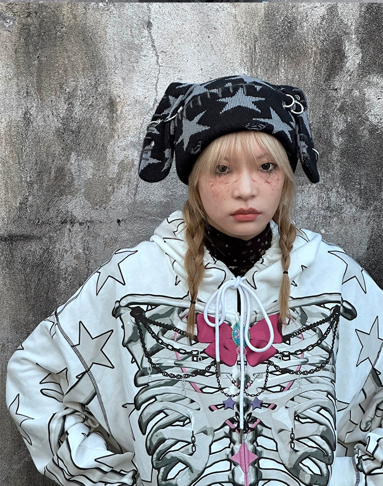 A blonde with braided hair and a neutral expression stands against a textured wall, embodying alternative fashion. They're wearing floppy-eared hat, polka dot turtleneck, and Seakoff's Gothic Skeleton Star Hoodie—full-zip with skeleton print and star pattern.