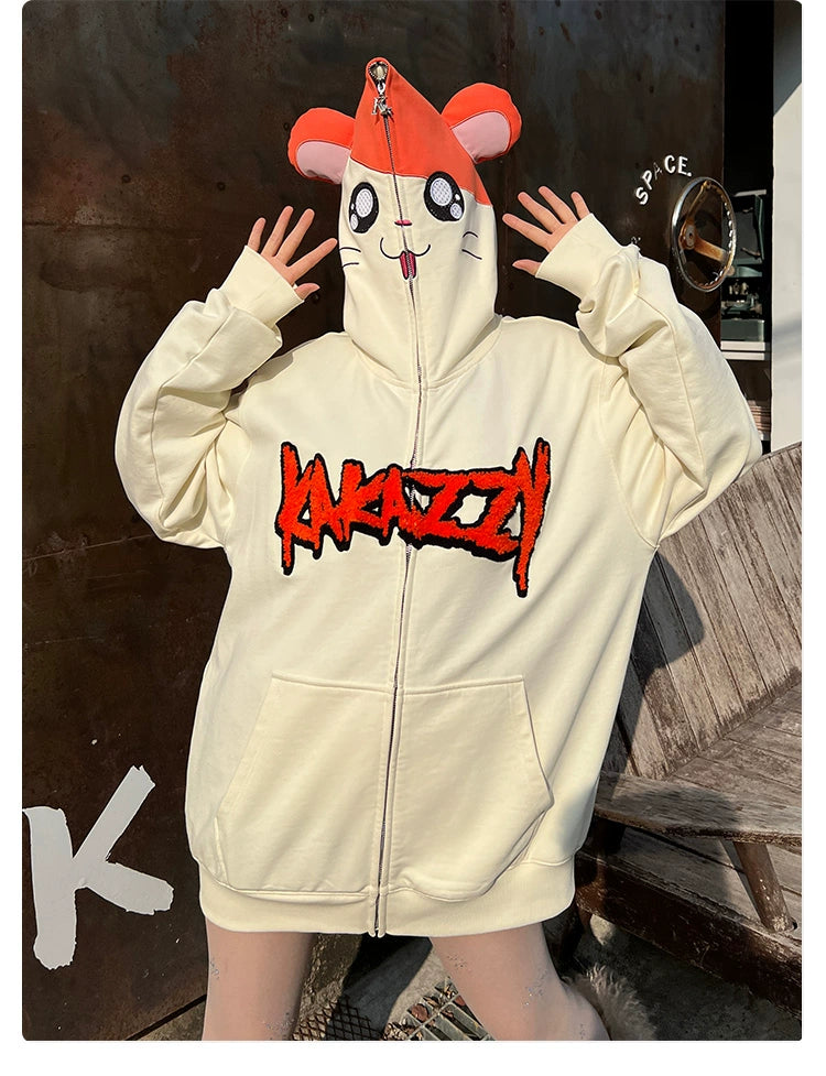 A person poses with hands up, wearing a Seakoff Cute Hamster Cartoon Hoodie crafted from 400GSM cotton featuring a cartoon face and playful ears on the hood. Its light-colored design with bold red text makes it a perfect gift for cartoon lovers against a weathered wall backdrop!.