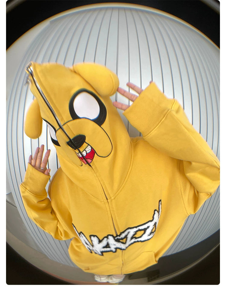 A person wears the Seakoff Kawaii Yellow Dog Hoodie, a 400GSM cotton oversized zip-up featuring ears and a playful dog face. The anime streetwear piece includes a white graphic on the front as they pose with raised hands against a striped background.