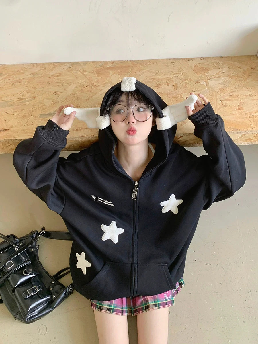 Seated on the floor against a wooden backdrop, a playful person in Seakoff's Kawaii Star Elf Hoodie—featuring 3D ears and soft star appliques—tugs at its strings. Beside them is a black bag with buckles, and their expression is delightfully playful.