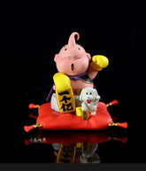 The Seakoff Majin Buu Collectible Figure is an 8.5cm Dragon Ball Z statue featuring the pink character with a single antenna and yellow gloves, sitting on a red cushion, holding a yellow object with black markings. A small white dog rests beside it against a black background. Lucky New Year Edition.