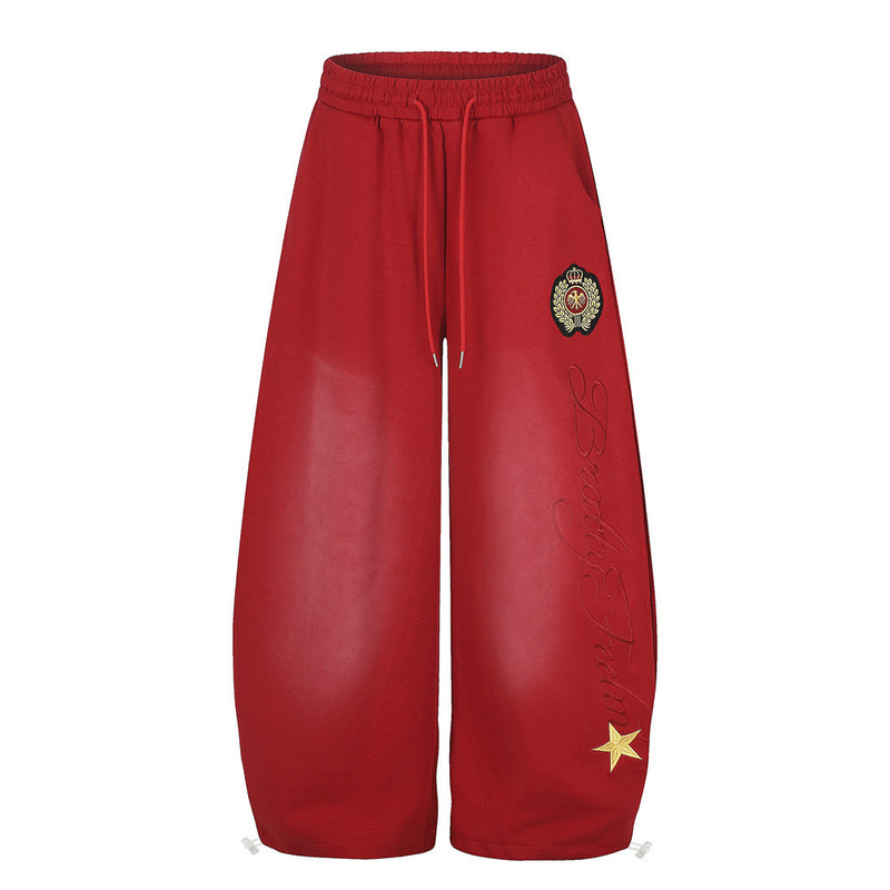 Men's oversized wide-leg sweatpants in red with cursive script embroidery, woven crest badge, and gold star appliqué