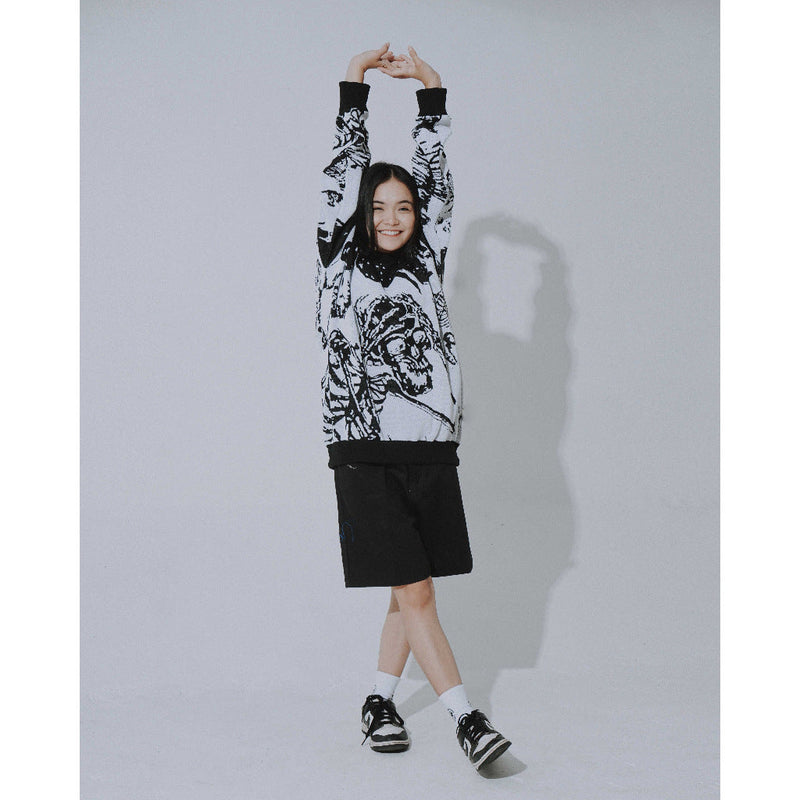 A person poses with arms raised, wearing Seakoff's Premium Skeleton Warrior Knit Sweater—a gothic jacquard design—paired with black shorts, sneakers, and white socks. Their shadow is cast on the light gray wall, enhancing the ensemble's gothic streetwear vibe.
