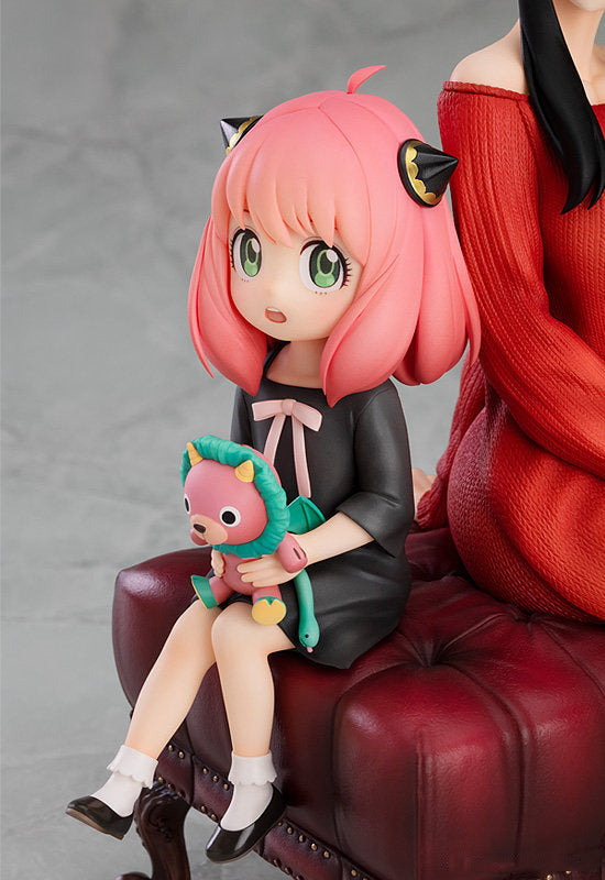 The Spy x Family Anya and Yor PVC Figure features a pink-haired girl in a black dress with cat ear headband, sitting on a red cushioned surface, large green eyes, white socks, and black shoes—ideal for any Spy x Family collection.