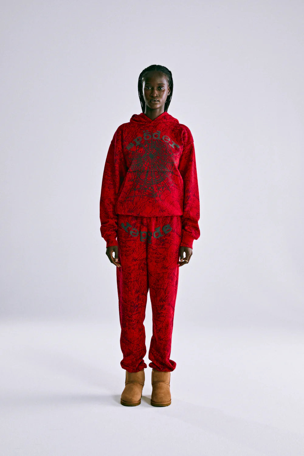 A person stands facing forward in a Red Sp5der Web Hoodie 440GSM – Red Spider Web Graphic Hoodie and matching sweatpants with a black abstract pattern, paired with light brown boots, against a plain light gray background.
