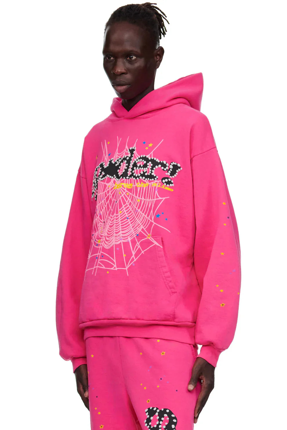Wearing the Sp5der “Nevermind the Punk” Hoodie in Hot Pink, featuring bold graphic designs like a white spider web and florals, this person stands sideways in vibrant 440GSM streetwear against a plain background.