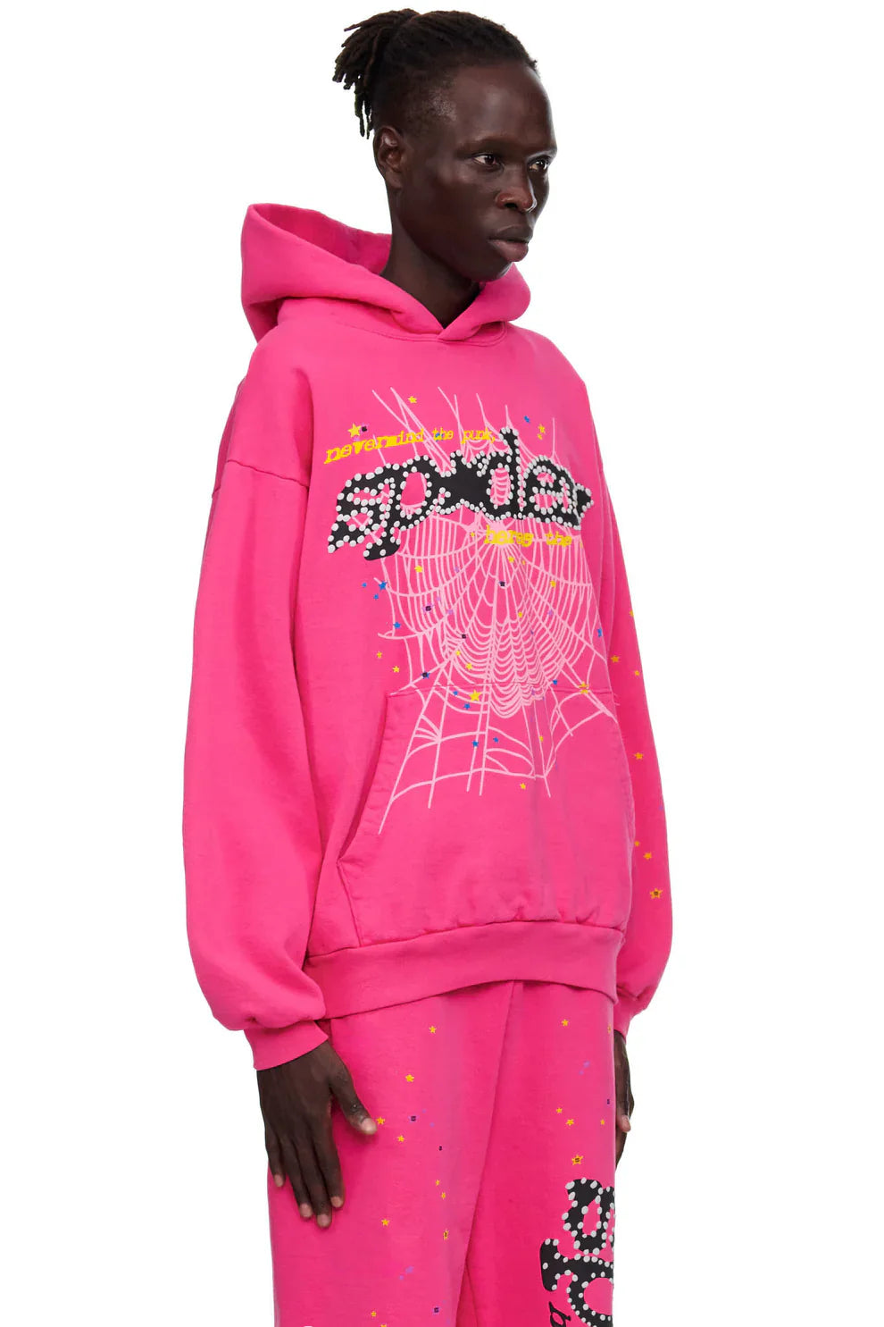 Someone wears the Sp5der “Nevermind the Punk” Hoodie in Hot Pink 440GSM and matching pants, both with spider web graphics and bold dotted "spider" text, accented by colorful confetti on a plain white backdrop.