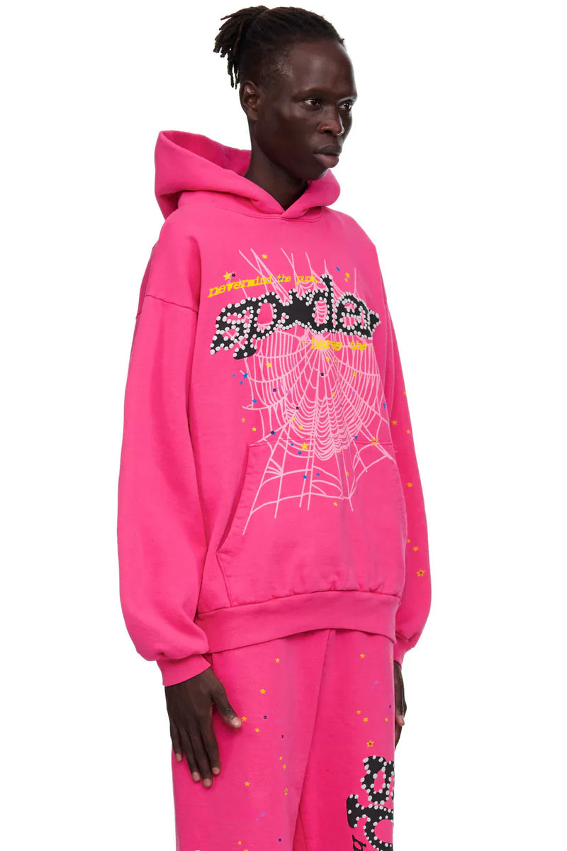 Someone wears the Sp5der “Nevermind the Punk” Hoodie in Hot Pink 440GSM and matching pants, both with spider web graphics and bold dotted "spider" text, accented by colorful confetti on a plain white backdrop.