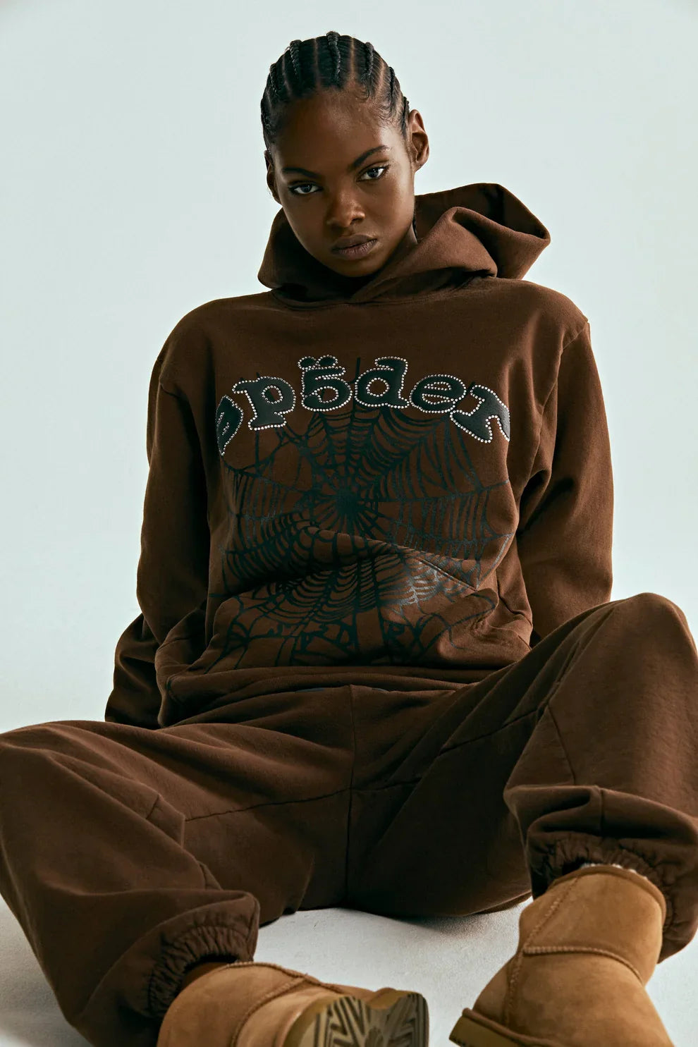 A person wears the Sp5der Web Hoodie – Mocha Brown 440GSM Heavyweight Streetwear Pullover with matching sweatpants and tan boots, sitting on the floor and looking at the camera against a plain background.
