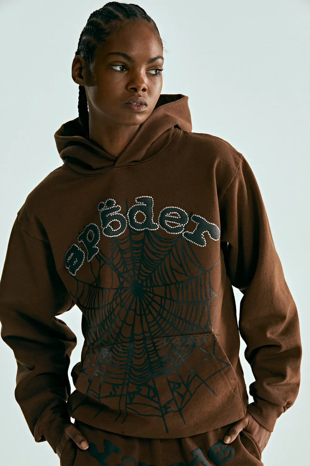 Someone wearing the Sp5der Web Hoodie – Mocha Brown 440GSM Heavyweight Streetwear Pullover, featuring a bold black spiderweb graphic and “spæder” across the chest, stands out with urban style against a plain light background.