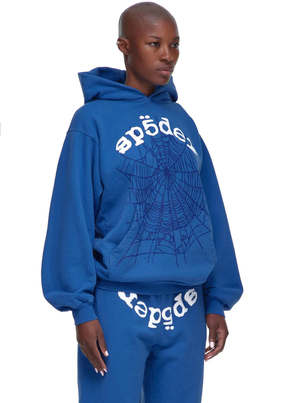 Wearing the Sp5der OG Web Hoodie 440GSM in royal blue, featuring bold spider-web graphics and white lettering on heavyweight cotton fleece, this person stands against a plain white background with matching sweatpants.