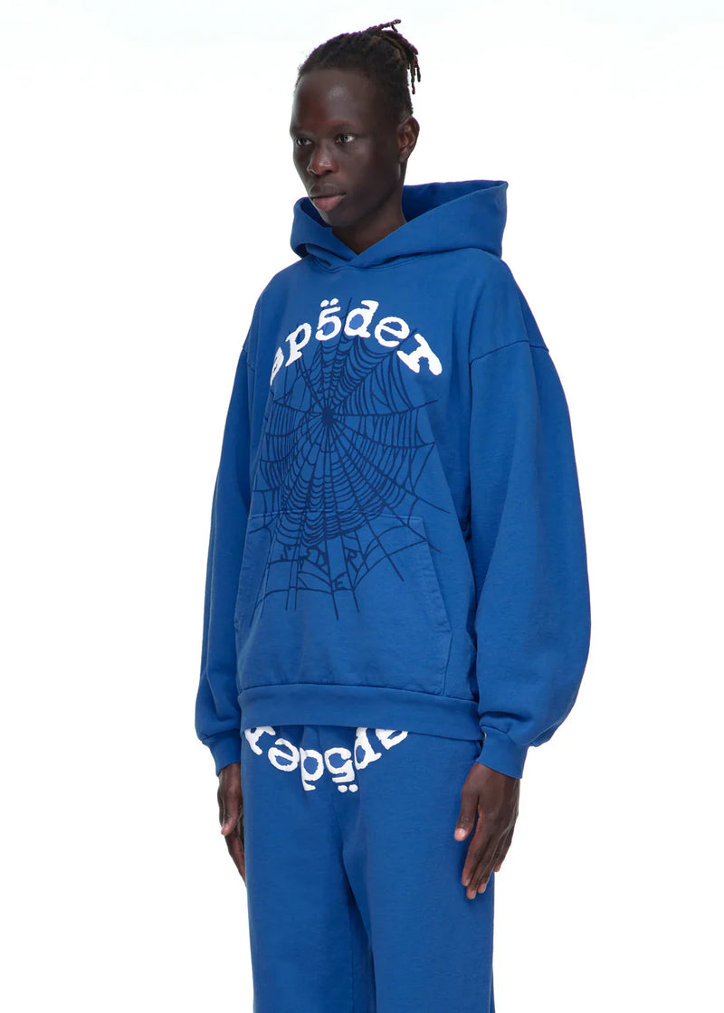 A person wears the Sp5der OG Web Hoodie 440GSM – Royal Blue Heavyweight Spider-Web Graphic Sweatshirt with matching blue sweatpants, standing against a plain white background.