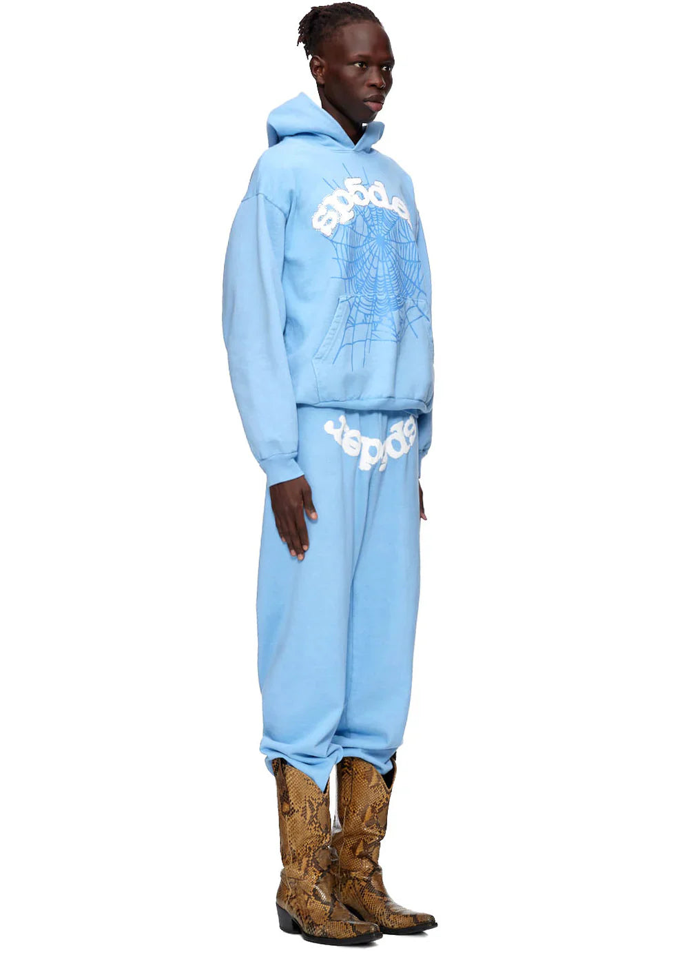 A person in profile models the Sp5der Hoodie – Sky Blue 440GSM Heavyweight Chenille Logo Spider-Web Streetwear Pullover with matching sweatpants and brown snakeskin cowboy boots, exuding street style against a plain white background.