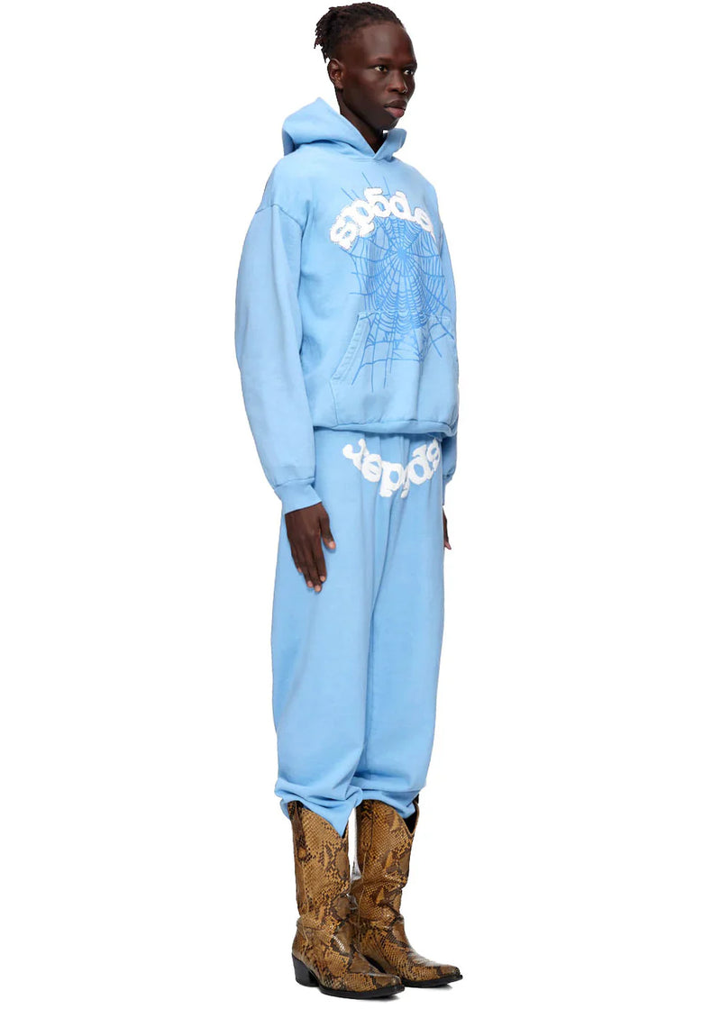 A person in profile models the Sp5der Hoodie – Sky Blue 440GSM Heavyweight Chenille Logo Spider-Web Streetwear Pullover with matching sweatpants and brown snakeskin cowboy boots, exuding street style against a plain white background.