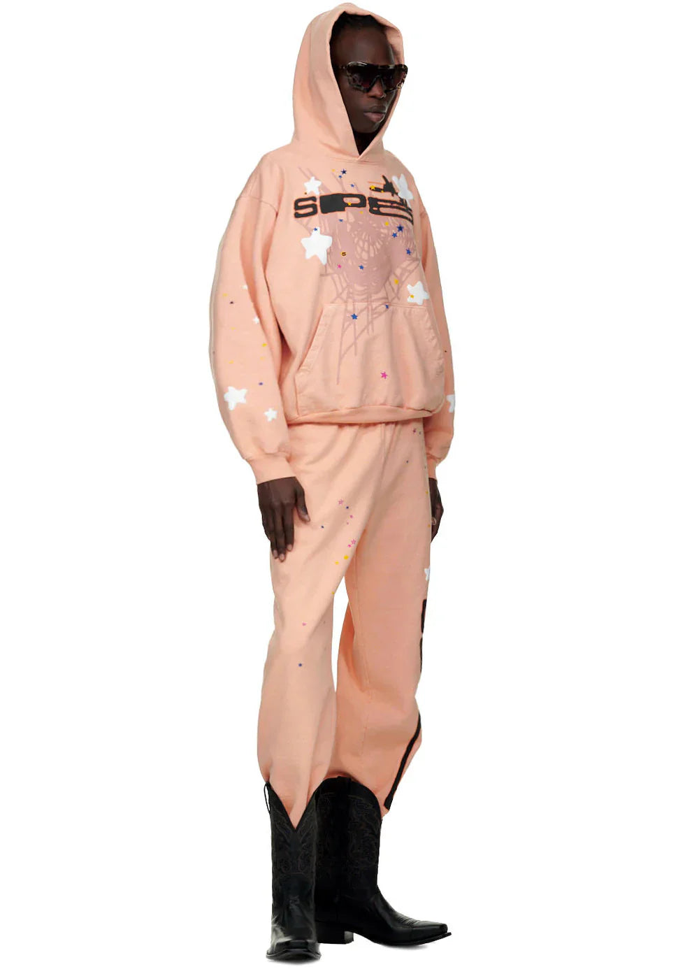 Wearing black sunglasses, matching pants, and black cowboy boots, a person stands sideways against a white background in a Sp5der Star Web Hoodie – Peach Pink 440GSM Heavyweight Oversized Streetwear Pullover, nailing bold Y2K vibes.