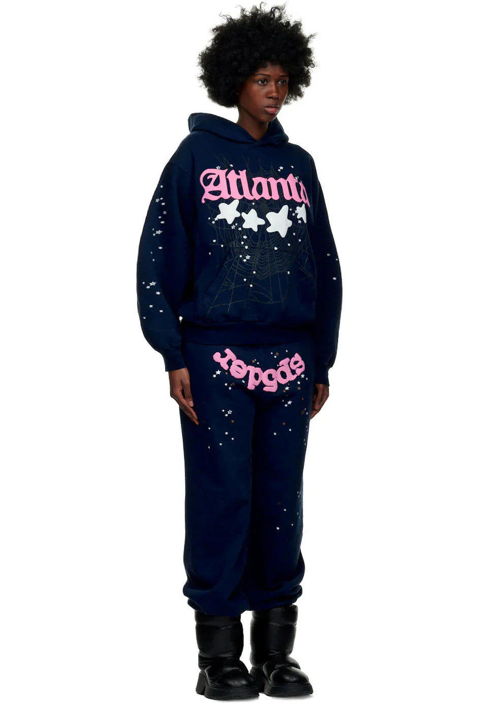 A person stands facing forward against a white background, wearing the navy blue Sp5der “Atlanta Nights” Web Hoodie (440GSM heavyweight oversized pullover) with matching sweatpants, featuring pink and white graphic text and splatter patterns, plus black boots.