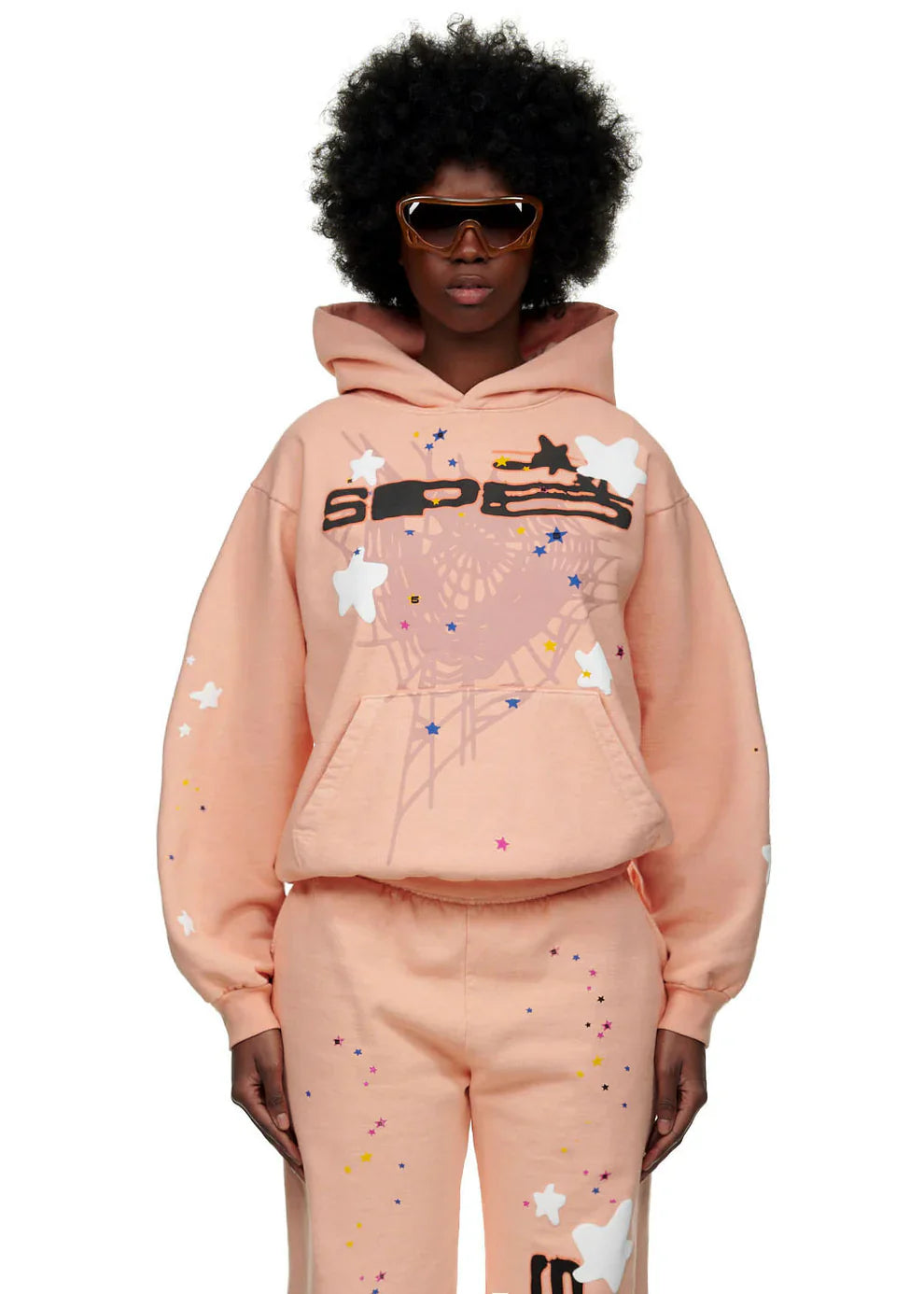 Wearing a Sp5der Star Web Hoodie – Peach Pink 440GSM Heavyweight Oversized Streetwear Pullover with matching sweatpants, star and paint splatter designs, and brown sunglasses, a person poses against a white background.