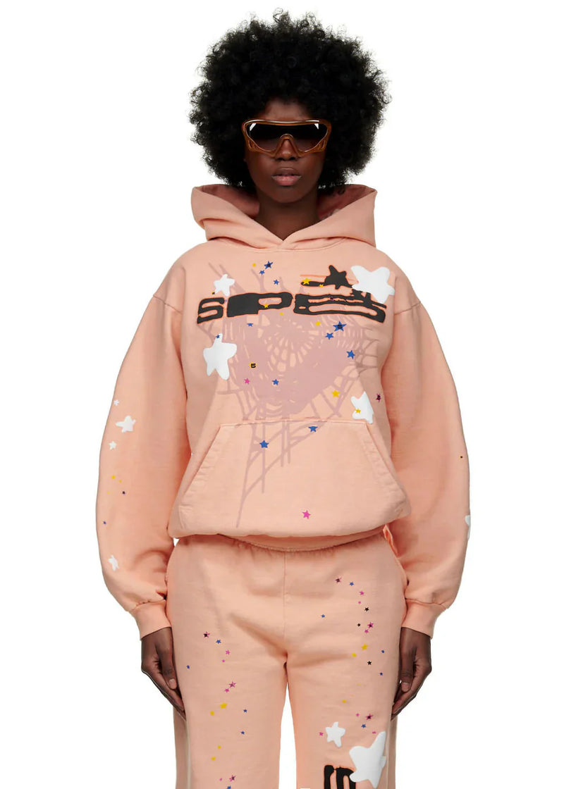 Wearing a Sp5der Star Web Hoodie – Peach Pink 440GSM Heavyweight Oversized Streetwear Pullover with matching sweatpants, star and paint splatter designs, and brown sunglasses, a person poses against a white background.