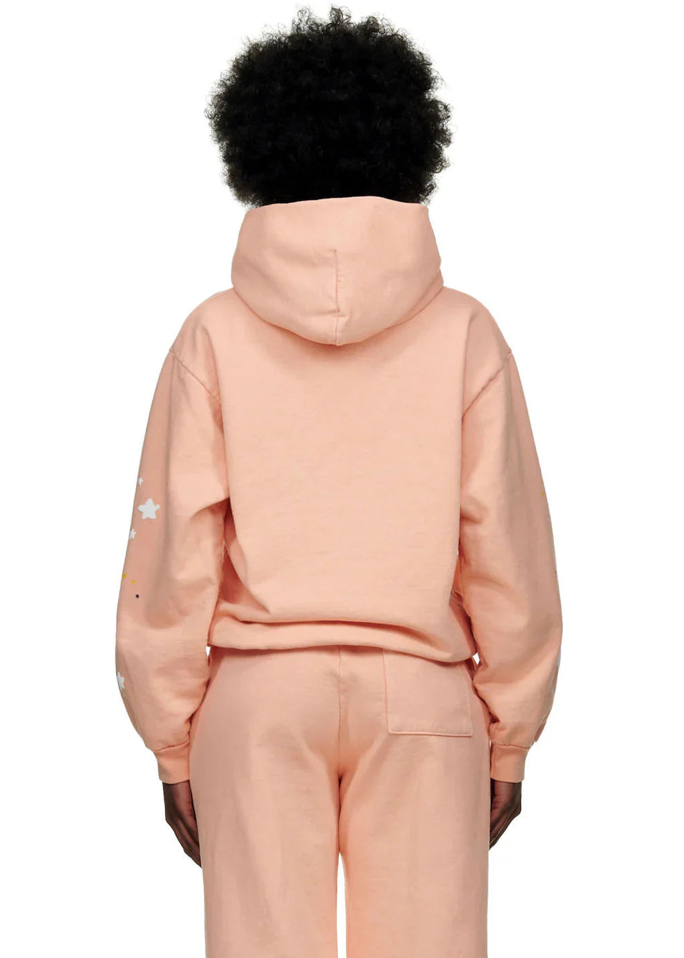 Person with short curly hair wears the Sp5der Star Web Hoodie – Peach Pink 440GSM Heavyweight Oversized Streetwear Pullover and matching sweatpants, hood up, with small star graphics on one sleeve—a Y2K-inspired look shown from the back.