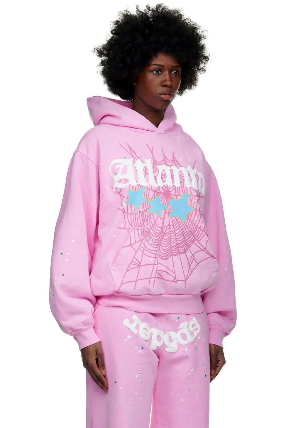 A person with short curly hair wears the Sp5der Atlanta Hoodie – Pink 440GSM Heavyweight Y2K Streetwear Pullover, featuring a white spiderweb, blue graphic, bold white text, and matching sweatpants with stylized lettering.