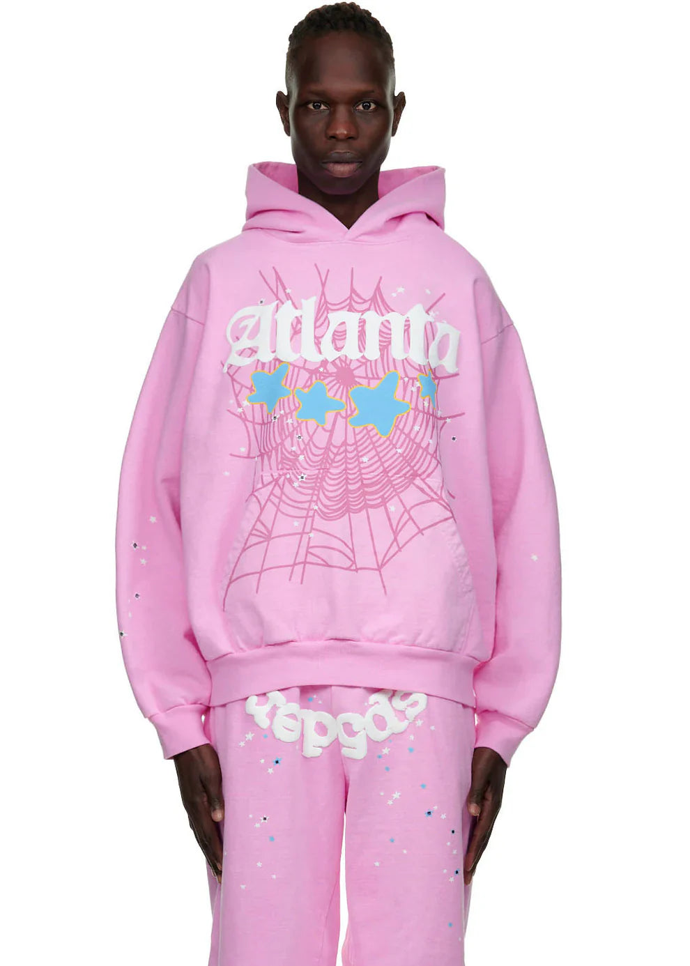 A person models the Sp5der Atlanta Hoodie – Pink 440GSM Heavyweight Y2K Streetwear Pullover, featuring a white web design, blue and yellow stars, and text, paired with matching pants against a plain background.