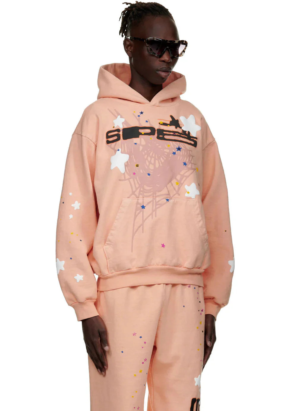 A person in dark sunglasses and matching pants wears the Sp5der Star Web Hoodie – Peach Pink 440GSM Heavyweight Oversized Streetwear Pullover with star and paint splatter designs, standing against a plain background.
