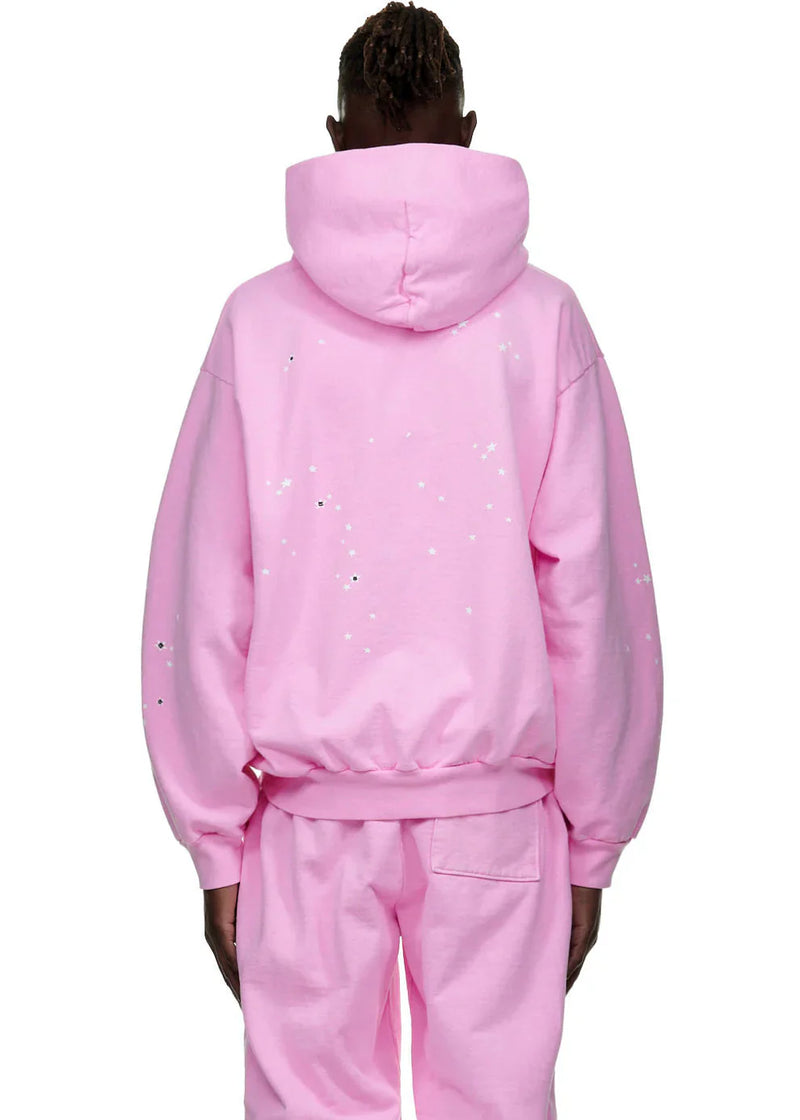 A person faces away from the camera in a Sp5der Atlanta Hoodie – Pink 440GSM Heavyweight Y2K Streetwear Pullover with matching pants. The hood is up, and the hoodie features white paint splatter details on the back and sleeves.