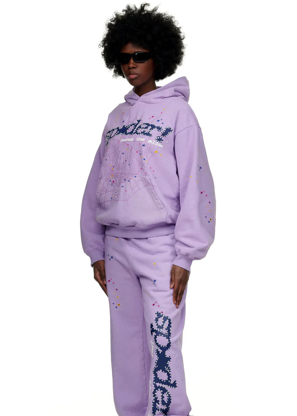 A person sports the Sp5der “Hares the Slime” lavender purple hoodie—440GSM heavyweight fleece with spider-web text and graphics—styled with matching sweatpants, an afro, black sunglasses, and vibrant streetwear vibes.