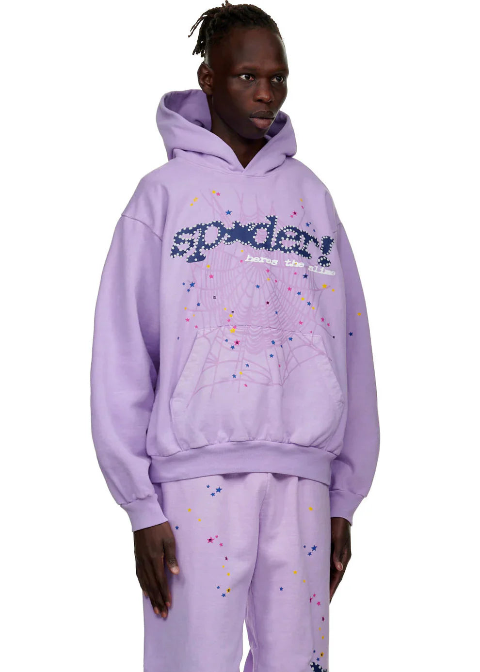 A person models the Sp5der “Hares the Slime” Hoodie in lavender purple, a 440GSM heavyweight pullover with a spider-web streetwear design, paired with matching pants and colorful speckles for a bold look against a plain white background.