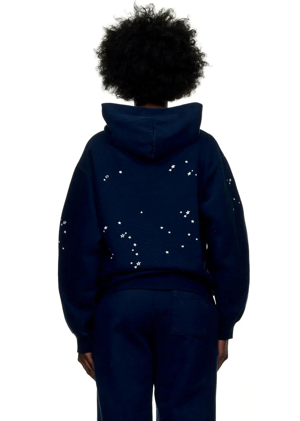 Individual with curly hair seen from behind in a navy blue Sp5der “Atlanta Nights” Web Hoodie—an oversized 440GSM heavyweight streetwear pullover with small white stars scattered on the back and sleeves.
