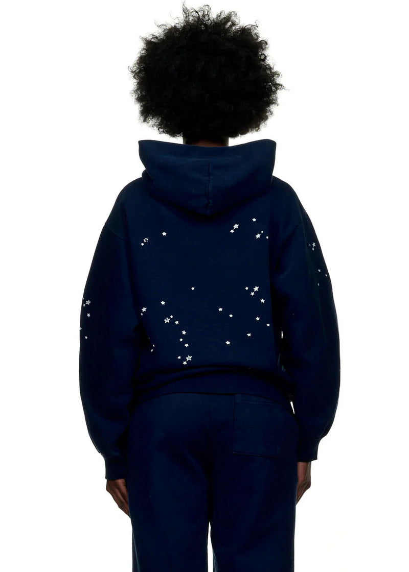 Individual with curly hair seen from behind in a navy blue Sp5der “Atlanta Nights” Web Hoodie—an oversized 440GSM heavyweight streetwear pullover with small white stars scattered on the back and sleeves.