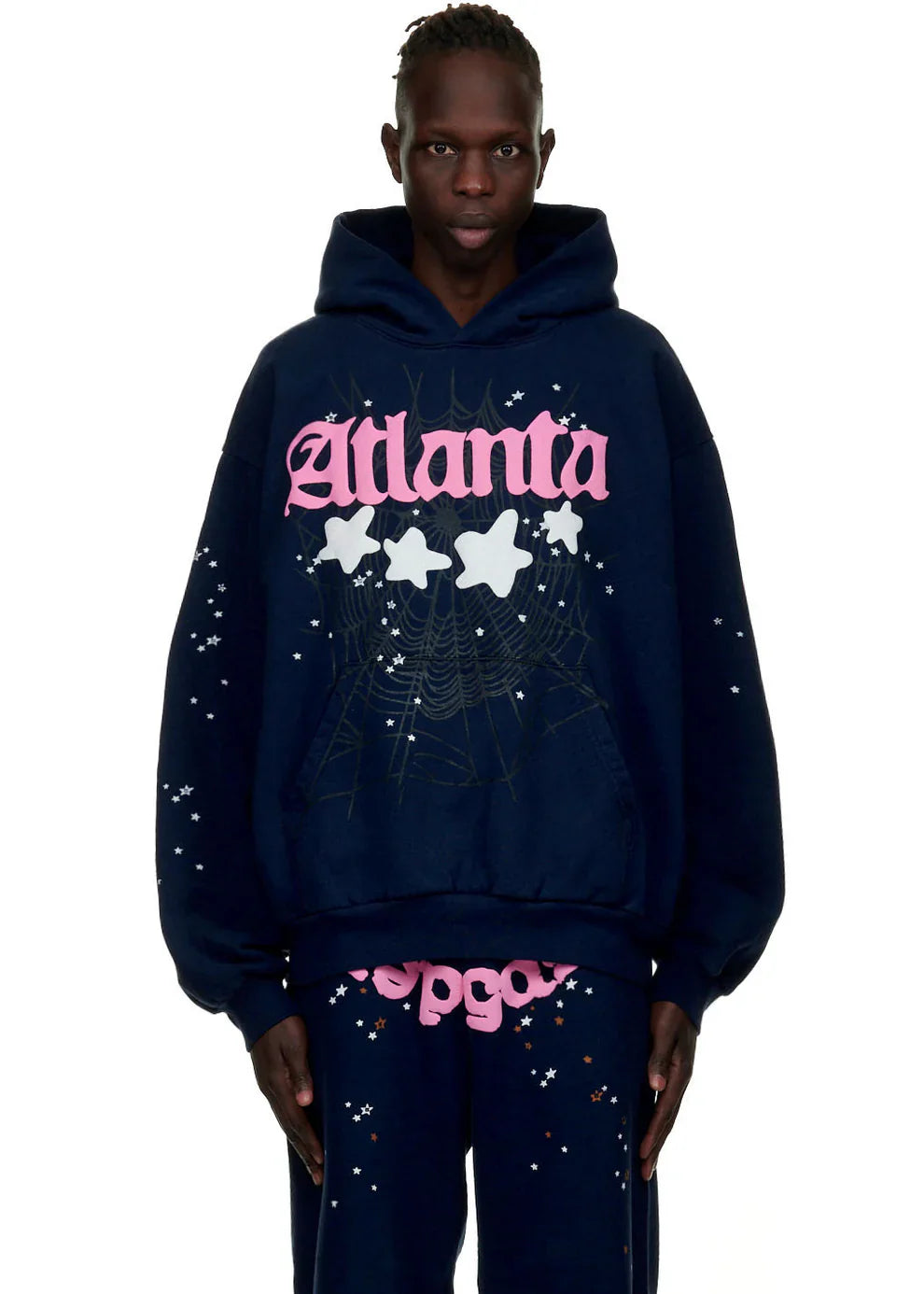 A person wears the Sp5der “Atlanta Nights” Web Hoodie – Navy Blue, a 440GSM heavyweight oversized streetwear pullover with pink text, white stars, and spiderweb graphic, paired with matching pants featuring white speckles and pink text.