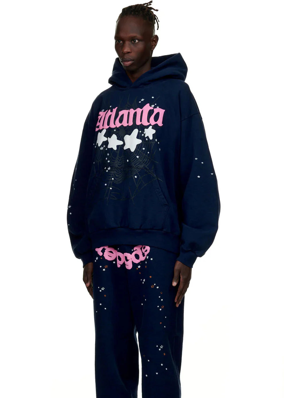 Wearing the Sp5der “Atlanta Nights” Navy Blue Heavyweight Hoodie and matching sweatpants, both with white paint splatters, pink ZILANIA text, white stars, a web graphic, and pink text on the waistband for a bold streetwear vibe.