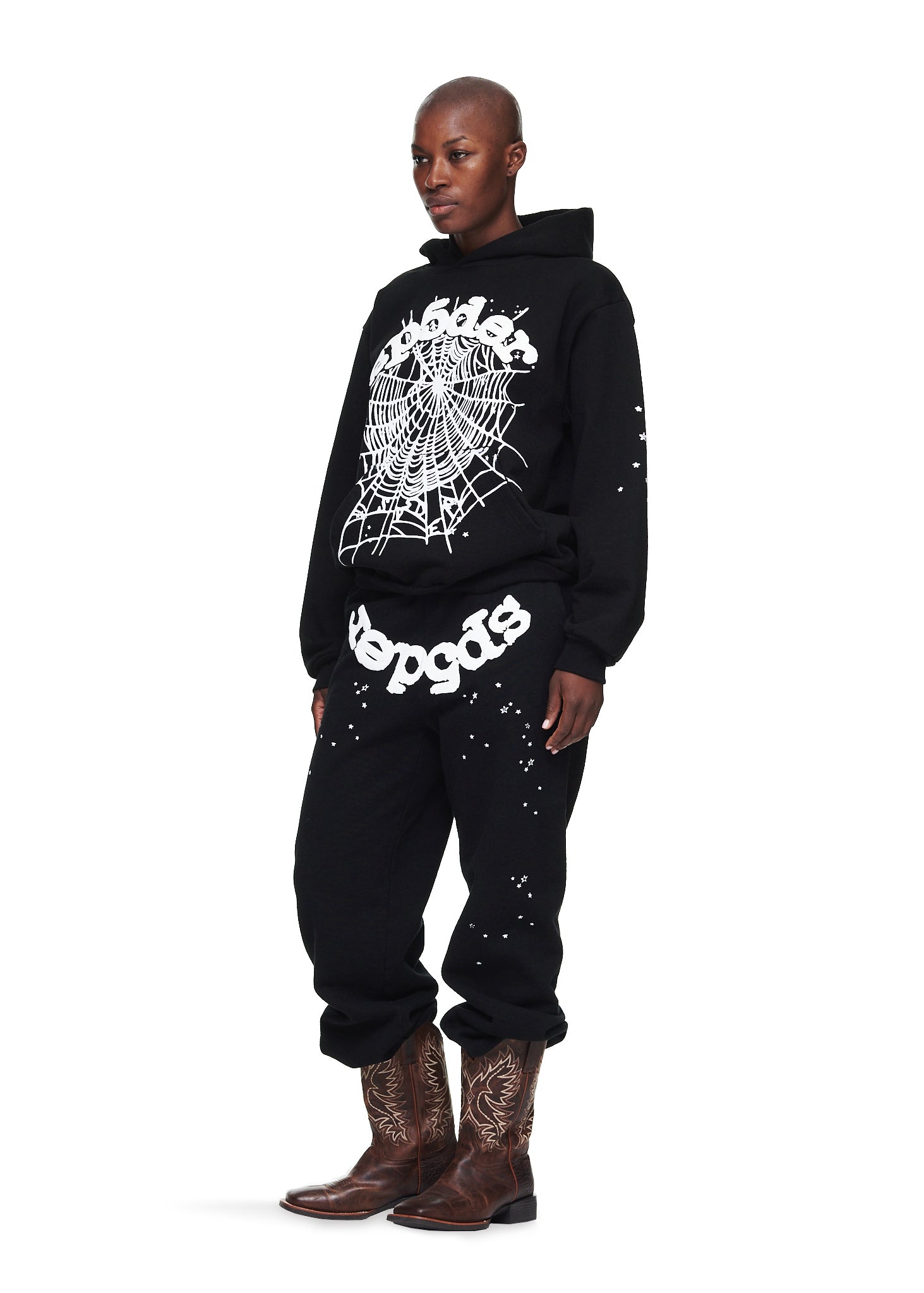 A person in a Sp5der “Webcore Noir” Hoodie – Black & White 440GSM Heavyweight Oversized Streetwear Pullover, matching black pants with white text, and brown embroidered cowboy boots stands against a white background.