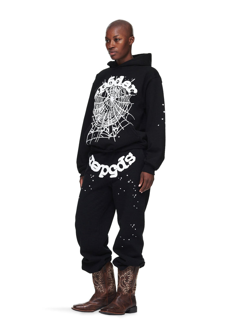 A person in a Sp5der “Webcore Noir” Hoodie – Black & White 440GSM Heavyweight Oversized Streetwear Pullover, matching black pants with white text, and brown embroidered cowboy boots stands against a white background.