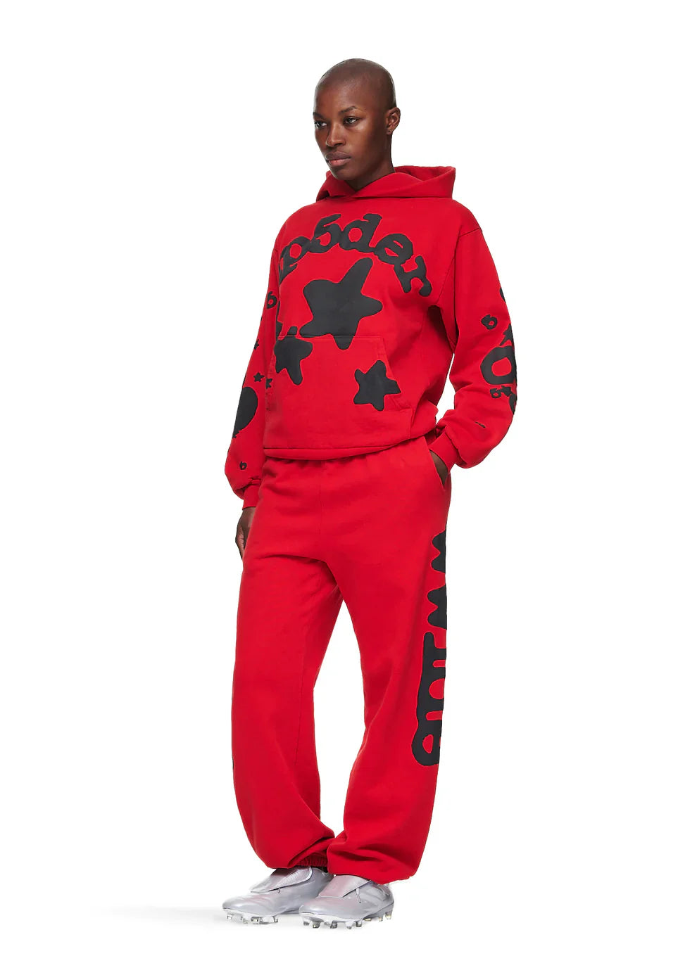 A person stands confidently in true Y2K streetwear style, wearing a red Sp5der “Starfire” Hoodie—an oversized 440GSM heavyweight pullover with black star graphics—paired with matching sweatpants and white sneakers.