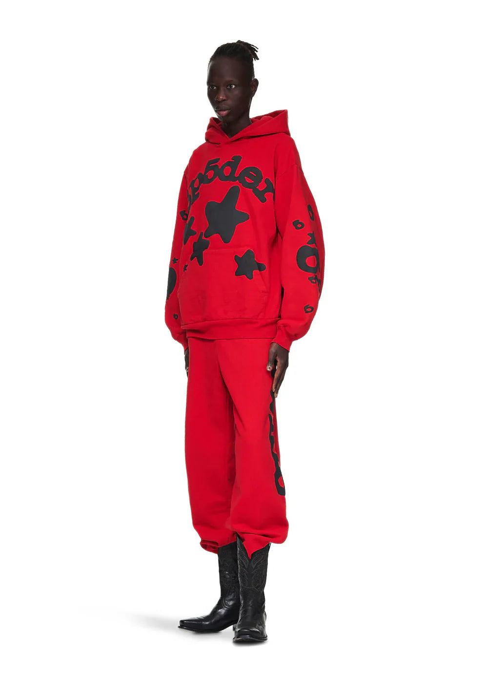 A person wears the Sp5der “Starfire” Hoodie – Red & Black 440GSM Heavyweight Oversized Streetwear Pullover with black star designs, matching red pants with black writing, and black cowboy boots, channeling Y2K style against a white background.