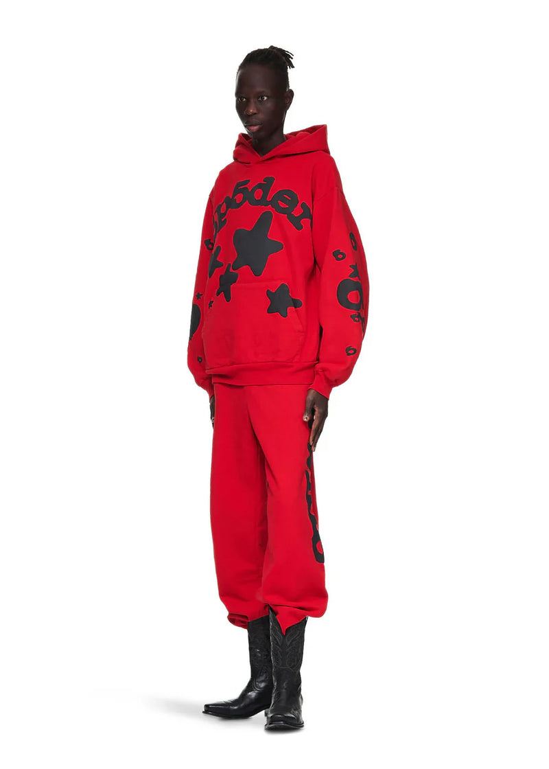 A person wears the Sp5der “Starfire” Hoodie – Red & Black 440GSM Heavyweight Oversized Streetwear Pullover with black star designs, matching red pants with black writing, and black cowboy boots, channeling Y2K style against a white background.