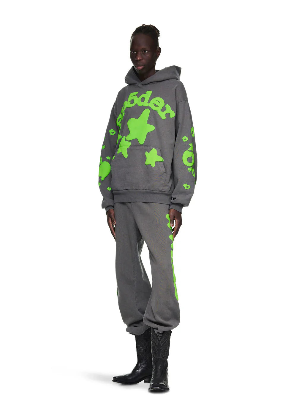 Wearing the Green and Grey Sp5der Hoodie – 440GSM Heavyweight Star Graphic Oversized Sweatshirt with bright green star designs, paired with sweatpants and black cowboy boots, a person stands against a white background, highlighting bold streetwear style.