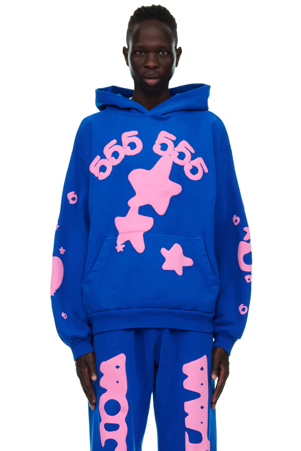 A person wears the Sp5der “555 Stars” Hoodie in royal blue/pink, a 440GSM heavyweight oversized streetwear pullover with bold pink numbers, stars, and abstract Y2K shapes, paired with matching pants for a vibrant look.