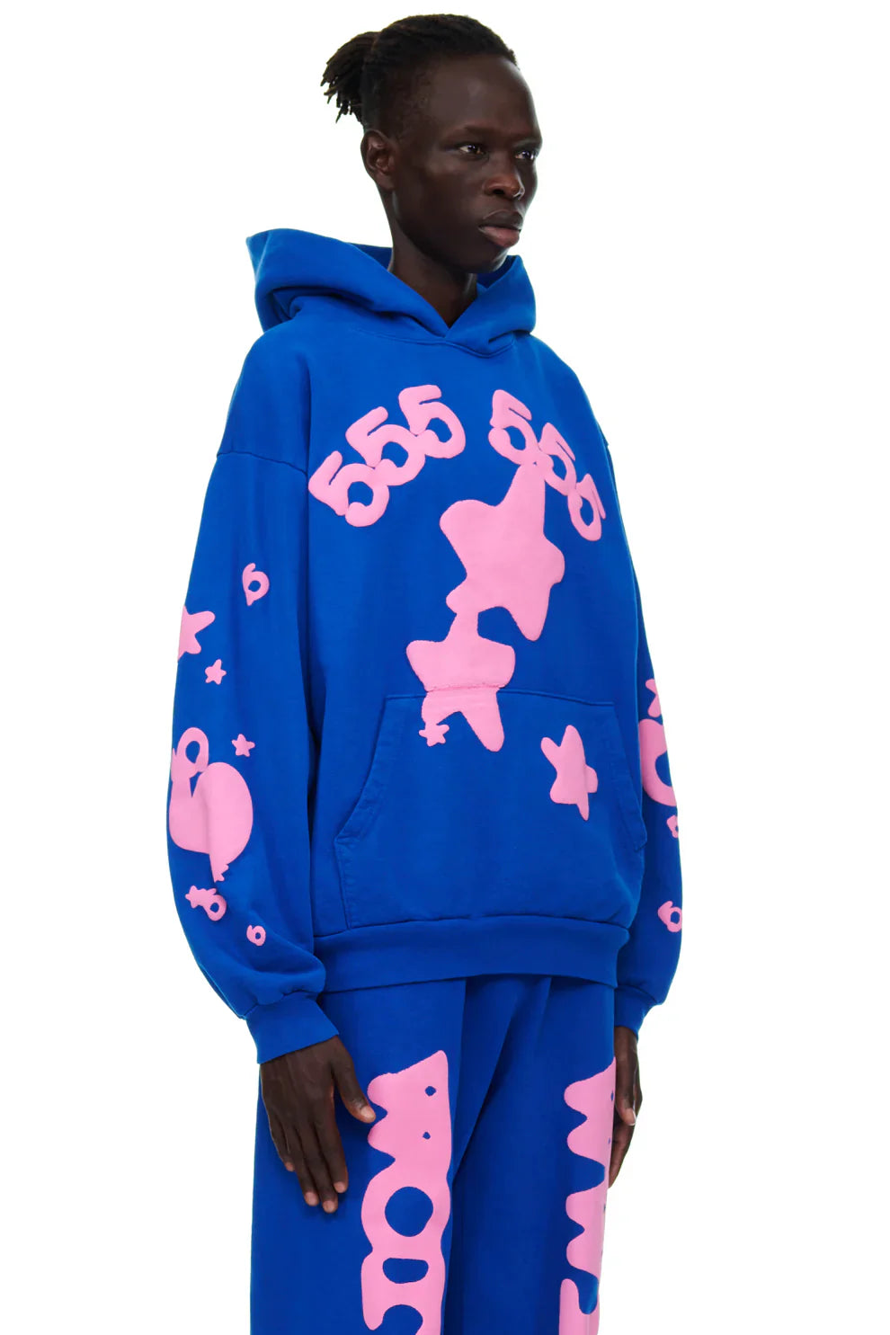 Someone wears the Sp5der “555 Stars” Hoodie in Royal Blue/Pink, a 440GSM heavyweight oversized streetwear pullover with matching pants featuring large pink abstract shapes and a front pocket, set against a white background.