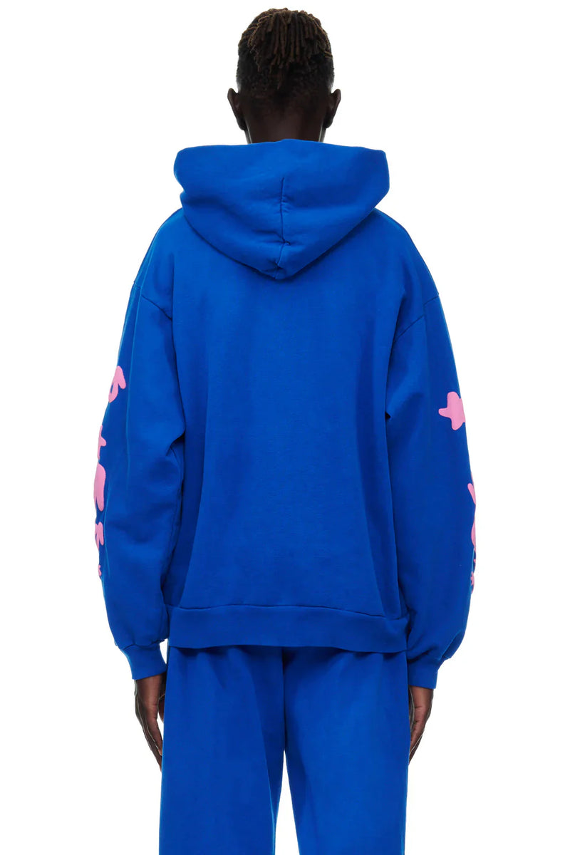 A person faces away wearing the Sp5der “555 Stars” Hoodie in Royal Blue/Pink, a 440GSM heavyweight oversized streetwear pullover, paired with matching blue pants, shown against a plain white background.