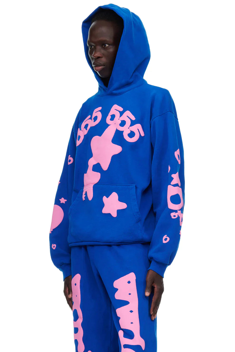 A person stands against a white background wearing the Sp5der “555 Stars” Hoodie in royal blue, featuring large pink graphic designs and numbers. This 440GSM heavyweight oversized streetwear pullover embodies bold Y2K style.