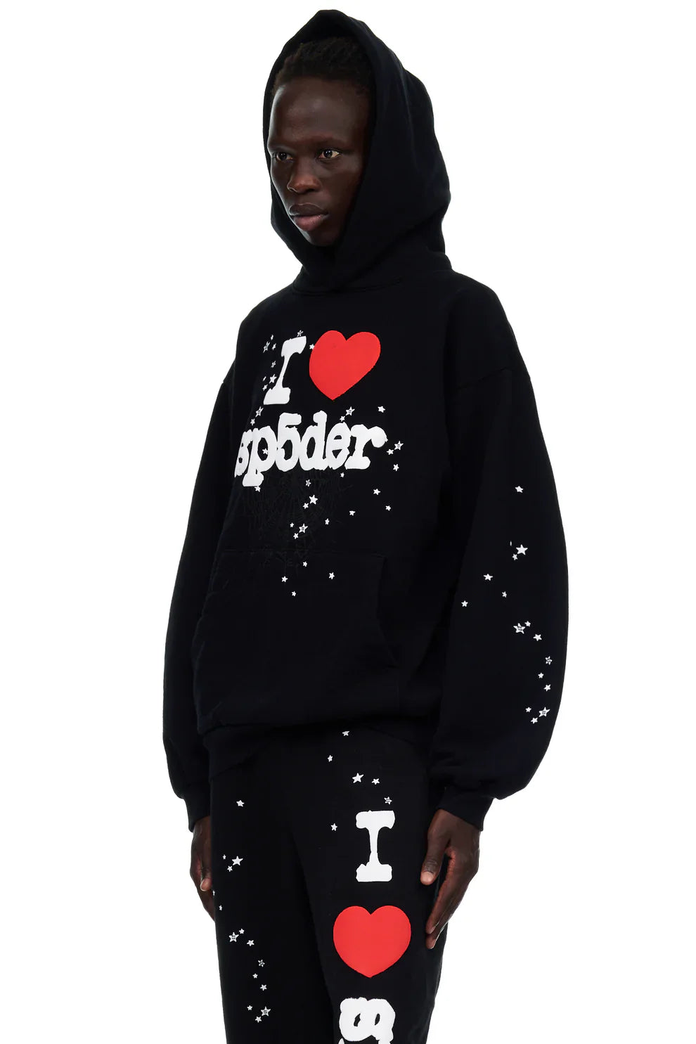 Wearing the Sp5der “I Love Sp5der” Hoodie – Black Web 440GSM Heavyweight Pullover with matching pants, white text, red heart graphics, and star speckles, the bold Y2K streetwear look stands out as the person faces slightly left.