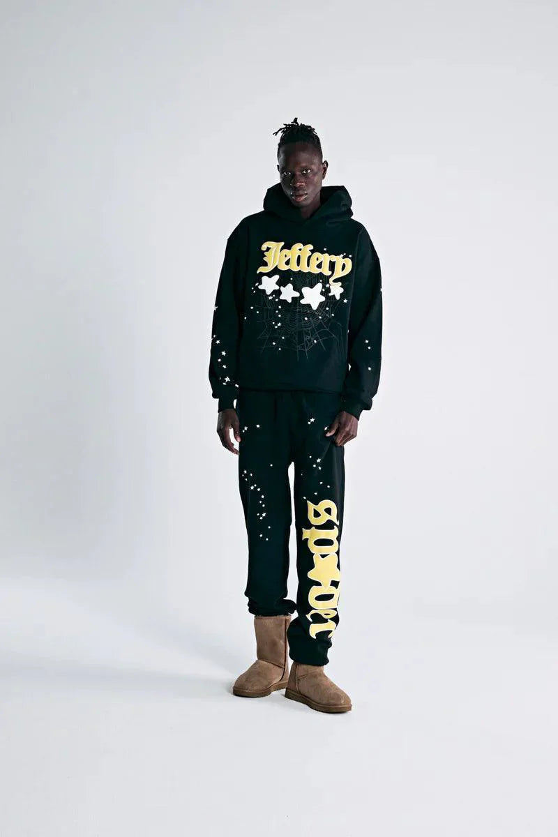A person stands against a plain background in the Sp5der Jeffery Hoodie – Black Web 440GSM Heavyweight Streetwear Pullover with matching sweatpants featuring bold yellow text, tan suede boots, and white paint splatters for a standout street style.