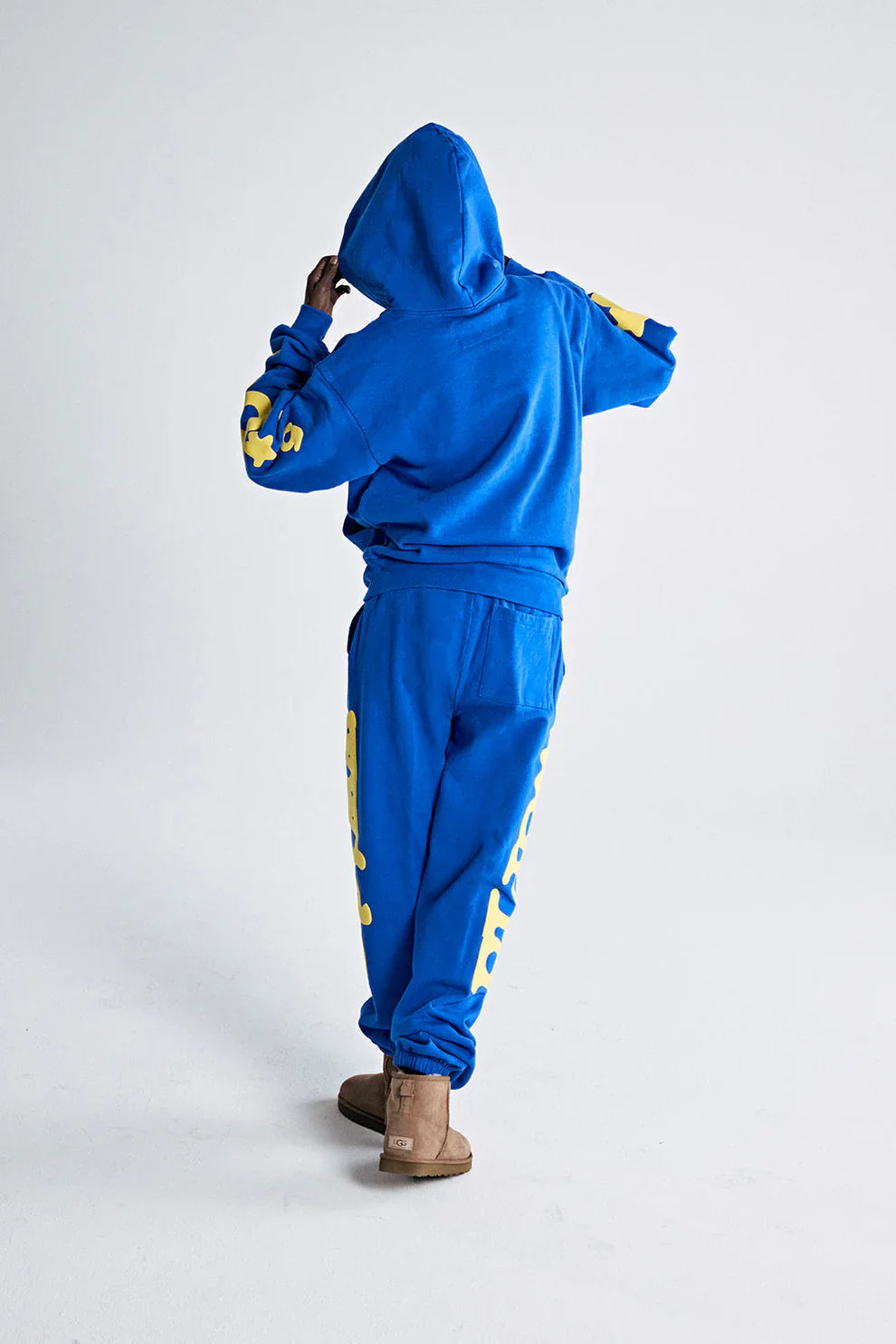 A person in the Sp5der 440GSM Heavyweight Hoodie – Royal Blue with Yellow Star 3D Puff Print stands with their back to the camera, adjusting their hood. They pair it with matching sweats and tan UGG-style boots against a plain white background.
